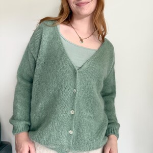 May include: A light green, button-down cardigan sweater. The sweater has a V-neck and is made of a soft, textured knit material. The buttons are a light, pearlescent color. The cardigan is worn over a light green scoop-neck top.
