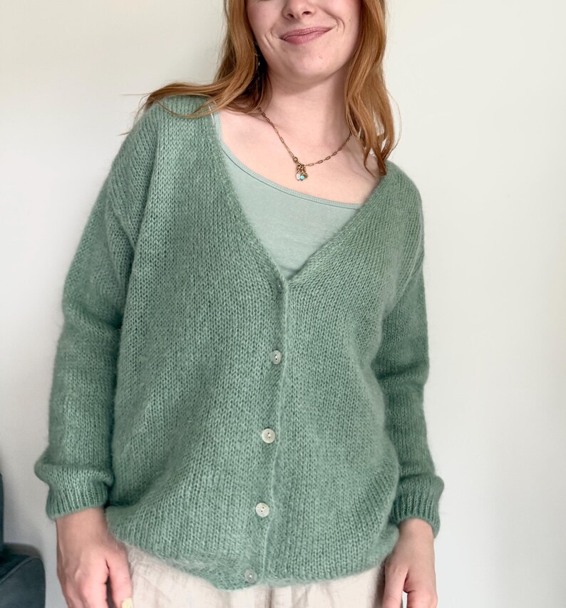 May include: A light green, button-down cardigan sweater. The sweater has a V-neck and is made of a soft, textured knit material. The buttons are a light, pearlescent color. The cardigan is worn over a light green scoop-neck top.