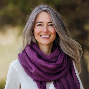 Puede incluir: Una mujer con cabello largo y gris sonríe, vistiendo una camisa blanca y una bufanda morada oscura. La bufanda está drapeada alrededor de su cuello y hombros, creando un efecto suave y fluido. El fondo está borroso, sugiriendo un entorno al aire libre.