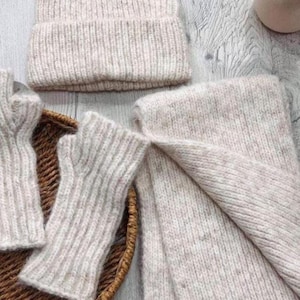 May include: A cozy, light beige knitwear set featuring a ribbed beanie, fingerless gloves, and a scarf. The items are arranged in a woven basket, showcasing the texture and warmth of the winter accessories. The set is perfect for cold weather.