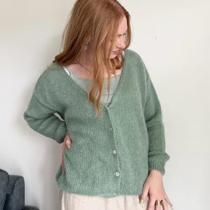 May include: A light green cardigan with a V-neck and button closure. The cardigan is made of a soft, textured knit fabric. The person is wearing a light green tank top and beige pants.