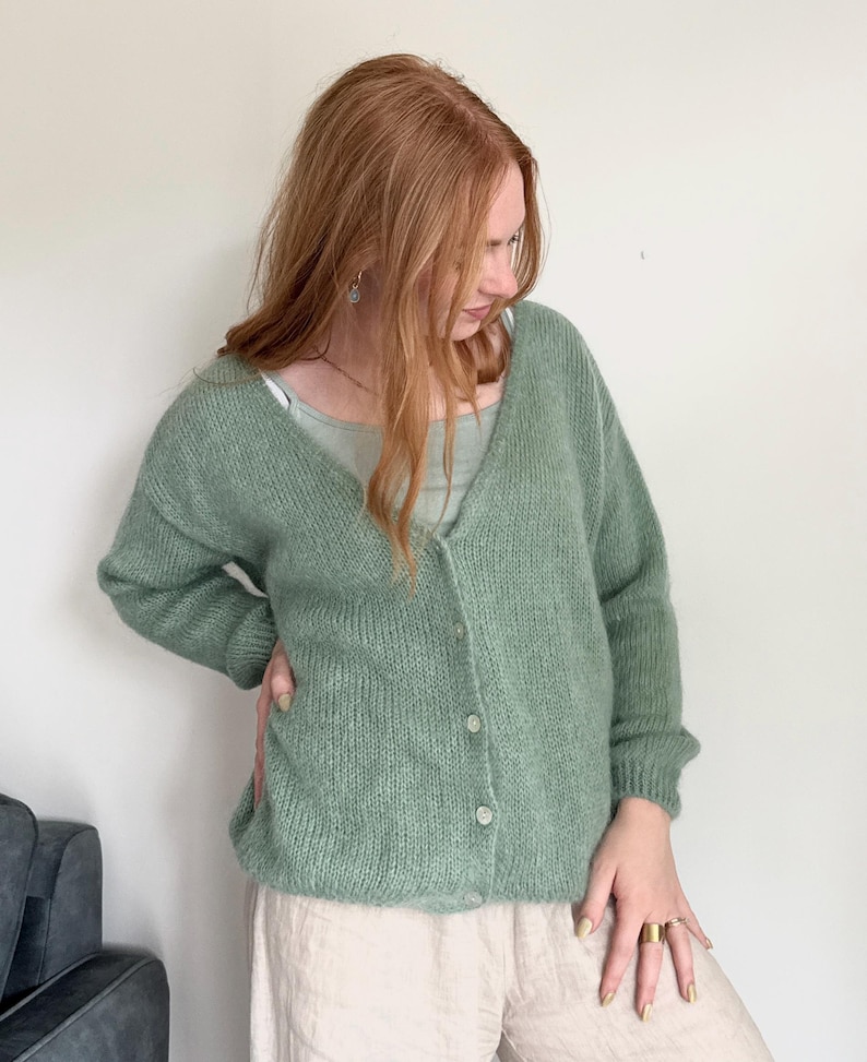 May include: A light green cardigan with a V-neck and button closure. The cardigan is made of a soft, textured knit fabric. The person is wearing a light green tank top and beige pants.