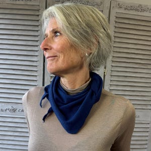 May include: A navy blue triangular scarf tied at the neck. The scarf is made of a soft, knitted material and is worn over a tan-colored turtleneck. The scarf is a versatile accessory.