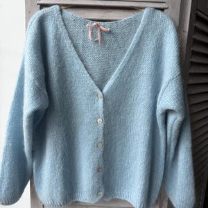 May include: A light blue, button-down cardigan sweater with a V-neck. The sweater has long sleeves and a soft, fuzzy texture. A small pink ribbon is tied at the neckline. The sweater is displayed against a light gray background.
