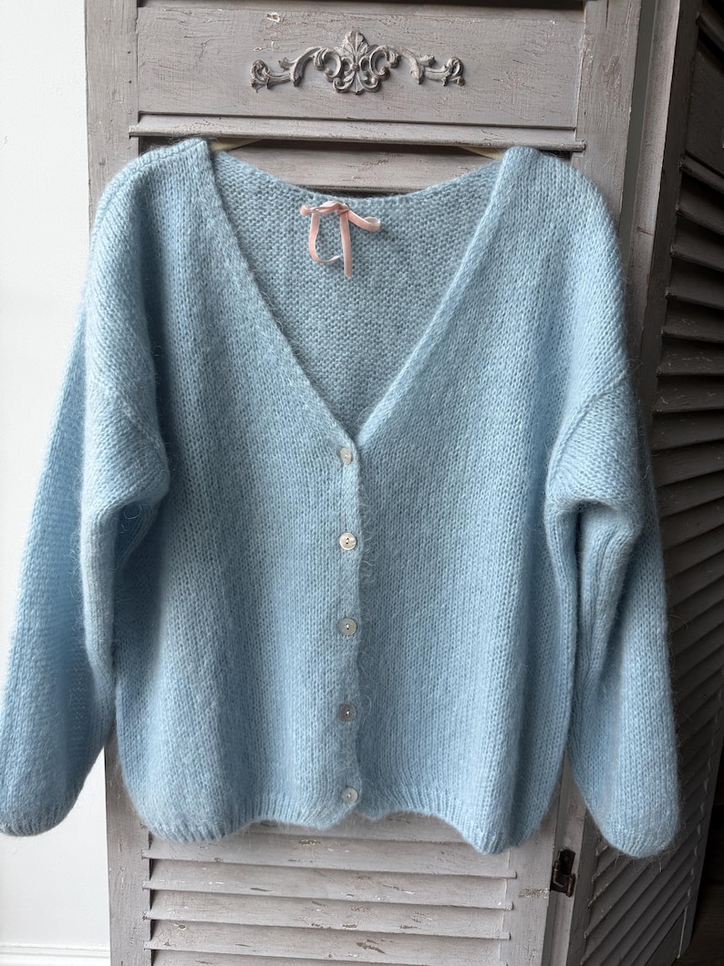 May include: A light blue, button-down cardigan sweater with a V-neck. The sweater has long sleeves and a soft, fuzzy texture. A small pink ribbon is tied at the neckline. The sweater is displayed against a light gray background.