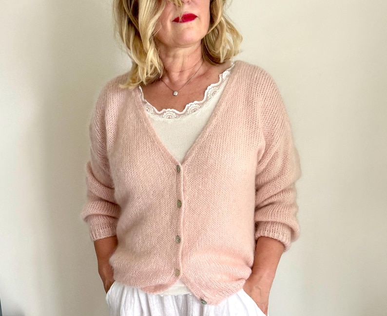 May include: A light pink, button-down cardigan with long sleeves. The cardigan is worn over a white top with lace trim. The buttons are a muted gold color. The cardigan is made of a soft, fuzzy material.
