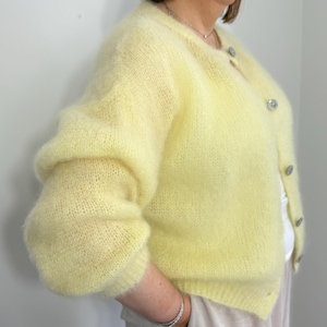 Puede incluir: Un cárdigan amarillo pálido con una textura difusa. El cárdigan tiene un cuello redondo y se abrocha con varios botones redondos plateados. Las mangas son largas y voluminosas. La prenda está hecha de un material suave y ligero.