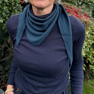Könnte beinhalten: Ein teal-farbener dreieckiger Schal über einem marineblauen Langarmshirt. Der Schal ist aus weichem Strickmaterial gefertigt. Die Person trägt ein goldenes Armband. Der Hintergrund zeigt grünes Laub.