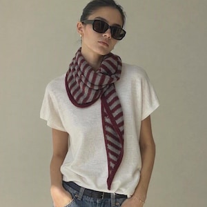 May include: A triangular knit scarf featuring a pattern of alternating dark red and grey stripes. The scarf is worn around the neck, paired with a plain off-white short-sleeved t-shirt, blue denim jeans with a black belt, and dark-framed sunglasses.