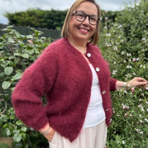 May include: A burgundy-colored, fuzzy cardigan with three white buttons. The cardigan is worn over a white top and paired with light-colored pants. The image is taken outdoors with green foliage in the background.