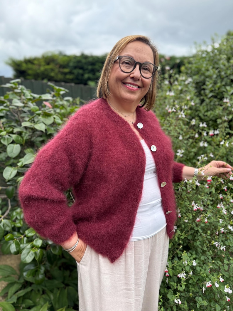 May include: A burgundy-colored, fuzzy cardigan with three white buttons. The cardigan is worn over a white top and paired with light-colored pants. The image is taken outdoors with green foliage in the background.