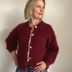 May include: A burgundy, fuzzy cardigan with a round neckline and large white buttons. The long-sleeved sweater is worn open, revealing a white top underneath. The woman in the image is wearing a gold necklace and a bracelet.