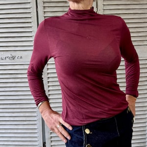 Puede incluir: Una camiseta de manga larga y cuello alto burdeos. La camisa está hecha de un material fino y semitransparente. La modelo lleva pantalones vaqueros azul oscuro con botones dorados.