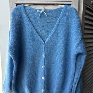 May include: A light blue, V-neck cardigan sweater with long sleeves. The sweater has a button-up front with five white buttons. The soft, fluffy knitwear is hanging on a wooden hanger with a blue ribbon.