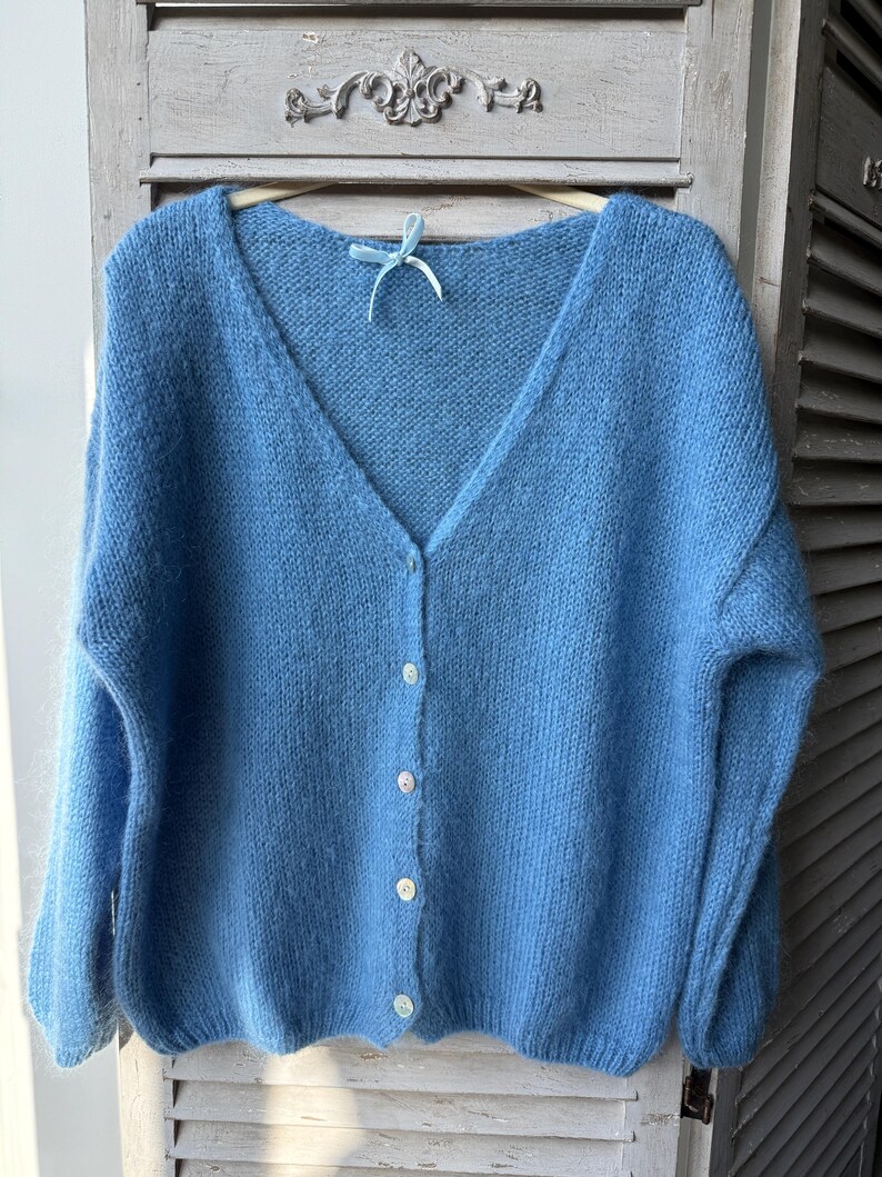 May include: A light blue, V-neck cardigan sweater with long sleeves. The sweater has a button-up front with five white buttons. The soft, fluffy knitwear is hanging on a wooden hanger with a blue ribbon.