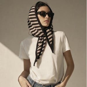 May include: A knitted headscarf featuring horizontal black and beige stripes, worn tied under the chin. The scarf is paired with a plain white short-sleeved t-shirt, blue denim jeans, and black rectangular sunglasses with dark lenses.
