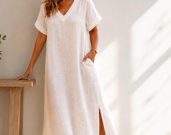 May include: A white midi-length dress made of a textured fabric, featuring a V-neckline, short sleeves with folded cuffs, and side pockets. The garment has a relaxed, loose-fitting silhouette that falls below the knees and includes a side slit.