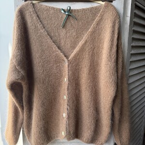 May include: A tan, V-neck cardigan sweater with long sleeves and a button-up front. The sweater is made of a soft, fuzzy material and hangs on a wooden hanger with a green ribbon.