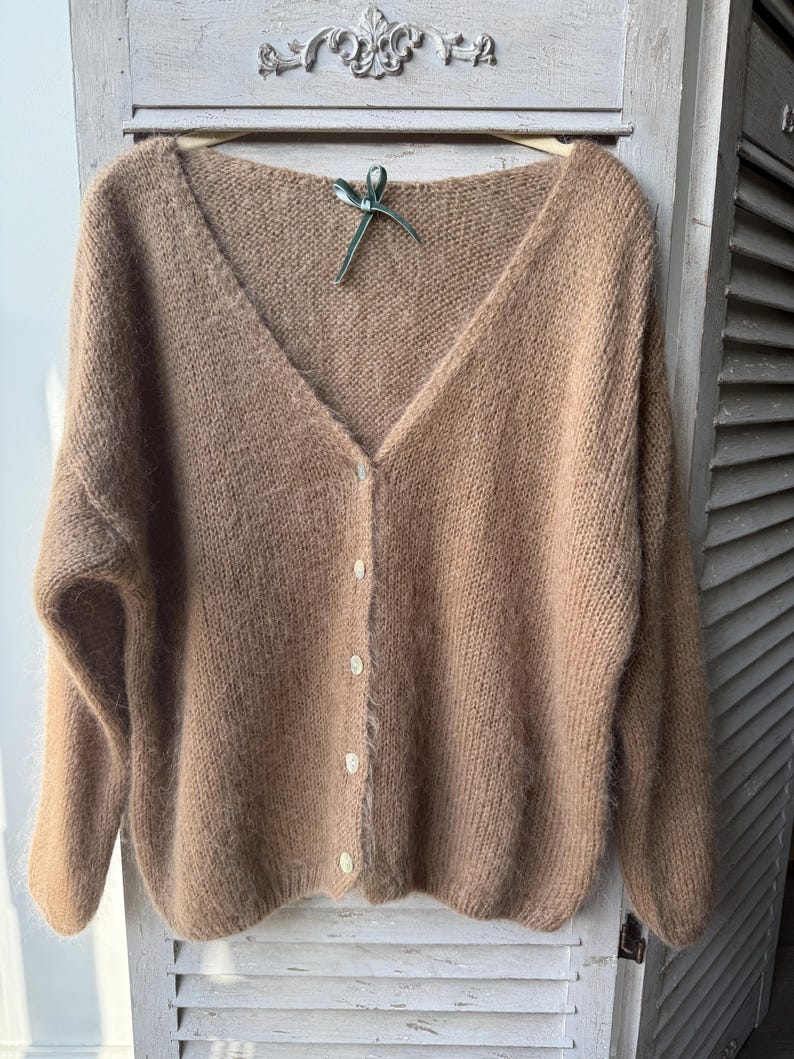 May include: A tan, V-neck cardigan sweater with long sleeves and a button-up front. The sweater is made of a soft, fuzzy material and hangs on a wooden hanger with a green ribbon.