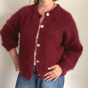 May include: A burgundy-colored, fuzzy cardigan with a round neckline and large, pearlescent buttons. The long-sleeved sweater is open and worn over a light-colored top and blue jeans. The cardigan has a soft, textured appearance.