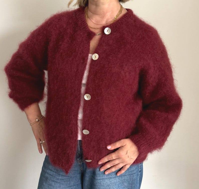 May include: A burgundy-colored, fuzzy cardigan with a round neckline and large, pearlescent buttons. The long-sleeved sweater is open and worn over a light-colored top and blue jeans. The cardigan has a soft, textured appearance.