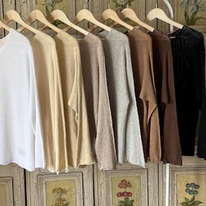 Op de afbeelding: Een collectie gebreide tops met lange mouwen in diverse neutrale kleuren, waaronder wit, beige, bruin en zwart, hangend aan houten kleerhangers. De tops hebben een los, vloeiend ontwerp, ideaal om te dragen.