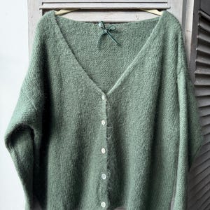 May include: A sage green, button-down cardigan sweater with a V-neck. The sweater is made of a soft, fuzzy material and has five white buttons down the front. A small green ribbon is tied at the neckline. The sweater is hanging on a wooden hanger.