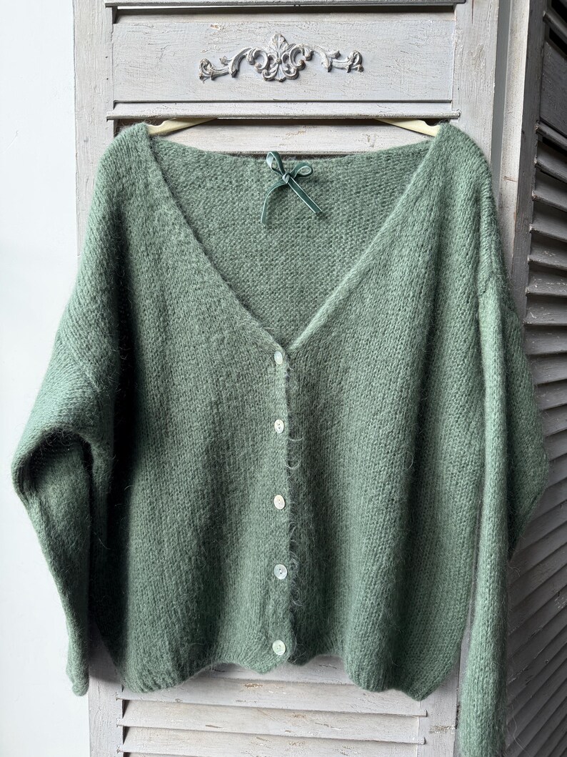 May include: A sage green, button-down cardigan sweater with a V-neck. The sweater is made of a soft, fuzzy material and has five white buttons down the front. A small green ribbon is tied at the neckline. The sweater is hanging on a wooden hanger.