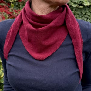 May include: A deep burgundy triangular scarf made of a fine-knit fabric. The scarf is draped around the neck, featuring a smooth, consistent texture and pointed ends that hang down over a dark navy blue long-sleeved shirt.