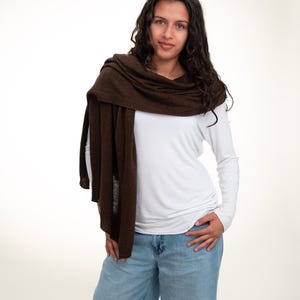 May include: A woman wearing a brown scarf draped over a white long-sleeve shirt and blue jeans. The scarf is a solid, dark brown color. The jeans are a light blue wash.