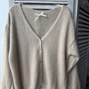 May include: A beige, button-down cardigan with a V-neck. The sweater is made of a soft, knitted material and has a relaxed fit. The cardigan is hanging on a wooden hanger. The buttons are a light, pearlescent color.