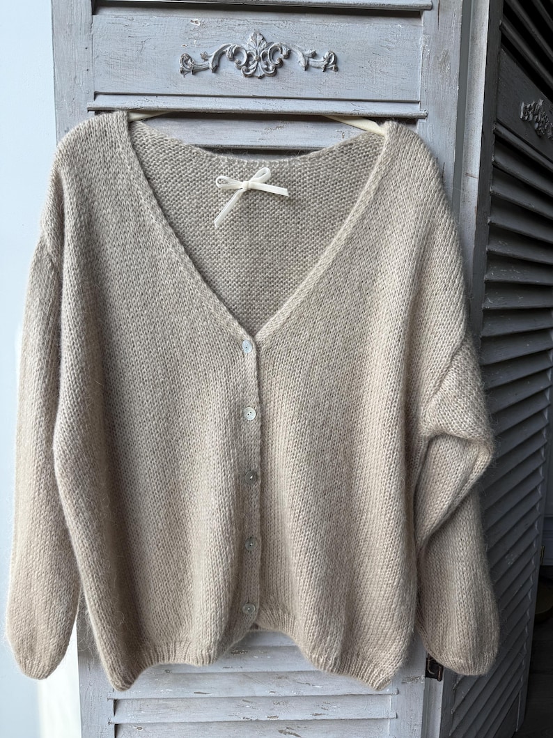 May include: A beige, button-down cardigan with a V-neck. The sweater is made of a soft, knitted material and has a relaxed fit. The cardigan is hanging on a wooden hanger. The buttons are a light, pearlescent color.