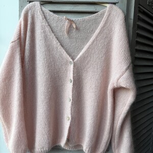May include: A light pink, oversized cardigan with a V-neck and button closure. The soft, fluffy knit sweater is hanging on a wooden hanger against a light gray background. The cardigan has long sleeves and a delicate, feminine design.