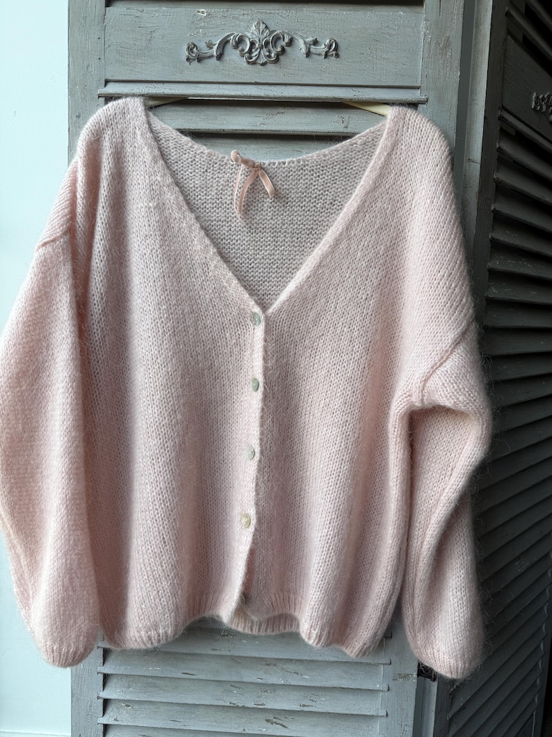 May include: A light pink, oversized cardigan with a V-neck and button closure. The soft, fluffy knit sweater is hanging on a wooden hanger against a light gray background. The cardigan has long sleeves and a delicate, feminine design.