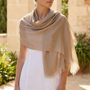 May include: A beige scarf draped over a white dress. The scarf has a fringed edge and is made of a soft, flowing fabric. The scarf is wrapped around the shoulders and neck.