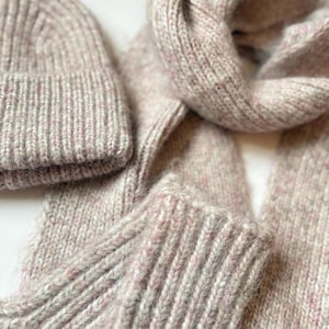 May include: A matching knitted hat and scarf set in a muted colour scheme of light grey and pink. The ribbed knit texture is prominent, suggesting warmth. The scarf is folded, and the hat is folded over.
