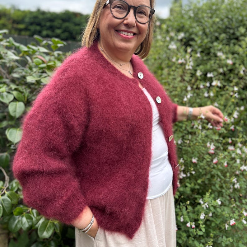 May include: A burgundy, fuzzy cardigan with three white buttons. The short-sleeved sweater is worn over a white top and tan skirt. The woman in the image is wearing glasses and smiling, with a green leafy background.