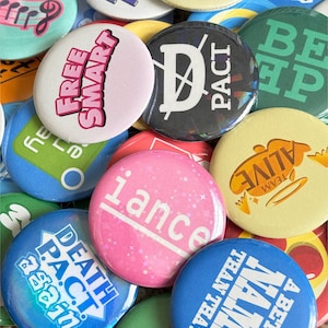 Puede incluir: Una colección de botones redondos y coloridos con varios diseños y texto. Algunos botones tienen frases como "FREE SMART" y "TEAM ALIVE". Otros botones presentan las palabras "PACT" e "iance".