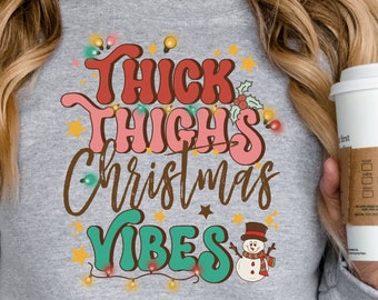 May include: A gray sweatshirt with the text "Thick Thighs Christmas Vibes" in a retro font with a snowman illustration. The text is surrounded by a string of colorful lights.