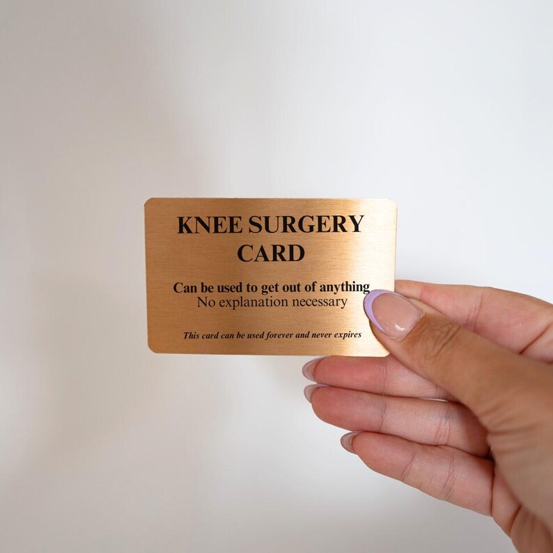 Op de afbeelding: Een rechthoekige, metallic goudkleurige kaart met zwarte tekst die luidt: KNEE SURGERY CARD, Can be used to get out of anything, No explanation necessary, This card can be used forever and never expires. De kaart heeft een geborstelde afwerking.