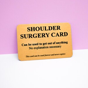 Op de afbeelding: Een rechthoekige goudkleurige metalen kaart met afgeronde hoeken en zwarte tekst die luidt: SHOULDER SURGERY CARD, Can be used to get out of anything, No explanation necessary, en This card can be used forever and never expires.