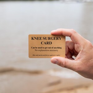 Op de afbeelding: Een rechthoekige goudkleurige metalen kaart met zwarte tekst: KNEE SURGERY CARD, Can be used to get out of anything, No explanation necessary, This card can be used forever and never expires. De kaart heeft een geborstelde afwerking.