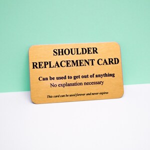 Op de afbeelding: Een rechthoekige goudkleurige metalen kaart met afgeronde hoeken en zwarte tekst die luidt: SHOULDER REPLACEMENT CARD, Can be used to get out of anything, No explanation necessary, en This card can be used forever and never expires.