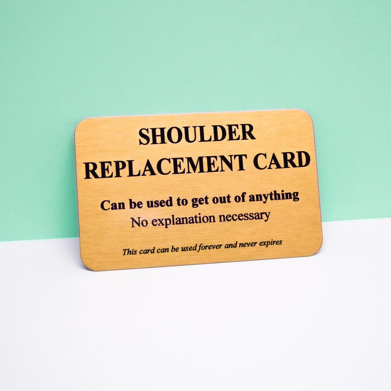 Op de afbeelding: Een rechthoekige goudkleurige metalen kaart met afgeronde hoeken en zwarte tekst die luidt: SHOULDER REPLACEMENT CARD, Can be used to get out of anything, No explanation necessary, en This card can be used forever and never expires.