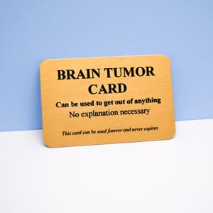 May include: A rectangular gold-coloured card with black text that reads: BRAIN TUMOR CARD, Can be used to get out of anything, No explanation necessary, This card can be used forever and never expires. The card has rounded corners.