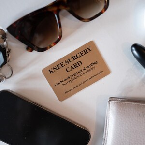 Op de afbeelding: Een rechthoekige goudkleurige metalen kaart met zwarte tekst die luidt KNEE SURGERY CARD, Can be used to get out of anything, No explanation necessary, en This card can be used forever and never expires, omringd door een zonnebril.