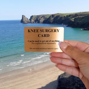 Op de afbeelding: Een rechthoekige goudkleurige metalen kaart met zwarte tekst: KNEE SURGERY CARD, Can be used to get out of anything, No explanation necessary, This card can be used forever and never expires. De kaart heeft een gladde, metalen afwerking.