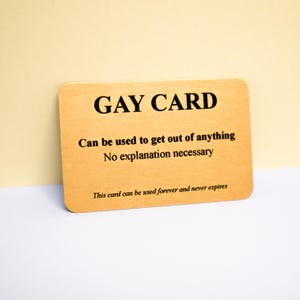 Peut inclure: Une carte rectangulaire en bois clair avec un texte noir indiquant GAY CARD, Can be used to get out of anything, No explanation necessary, et This card can be used forever and never expires. La carte présente des coins arrondis.