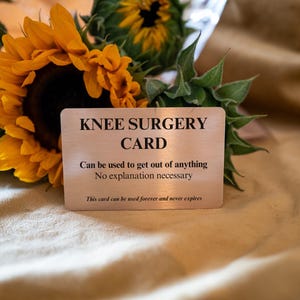 Op de afbeelding: Een rechthoekige zilverkleurige metalen kaart met zwarte tekst: KNEE SURGERY CARD, Can be used to get out of anything, No explanation necessary, This card can be used forever and never expires. De kaart is geplaatst voor zonnebloemen.