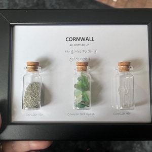May include: A framed display with three small glass bottles containing Cornish tin, sea glass, and air. The frame has a black border and a white background with the text "CORNWALL ALL BOTTLED UP Mr & Mrs Polding 09/09/2014" and the labels "Cornish Tin", "Cornish Sea Glass", and "Cornish Air".